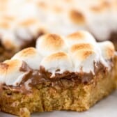 A close-up of a gooey dessert bar topped with toasted marshmallows and melted chocolate on a golden brown cookie base. The bar has a bite taken out of it, revealing its soft texture.