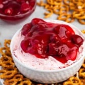 A white bowl filled with creamy pink Strawberry Pretzel Salad Dip topped with cherry sauce sits on a plate surrounded by mini pretzels. In the background, theres a small bowl of cherry sauce and a few scattered pretzels on a light-colored surface.